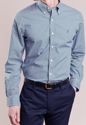 Chemise à carreaux verte et marine avec un col pointu, des manches longues et un petit logo sur la poitrine, associée à un pantalon foncé.