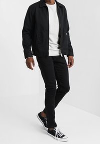 Chaqueta negra con cremallera sobre un sudadera gris claro, combinada con unos jeans skinny negros y zapatillas de lona negras. Diseño simple y casual.
