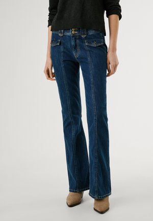 Tummansinisen bootcut-farkut edessä ompeleilla, nappiläpällisillä taskuilla, yhdistettynä mustaan pitkähihainen puseroon ja beigen värisiin kärjestä teräviin saappaisiin.