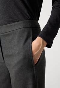 Pantalones de cuadros grises con una textura suave y una cinturilla ancha. Una mano está colocada en el bolsillo lateral, oscureciendo parcialmente el detalle.
