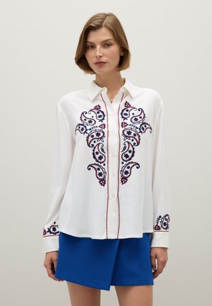 Camicia bianca con bottoni e ricamo paisley blu navy e rosso, con collo e polsini abbottonati. Abbinata a una gonna blu.