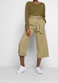 Persoon in een olijfgroene lange mouw top, beige hoge taille wijde cropped broek met knoopceintuur, en witte sneakers die tegen een witte achtergrond staat.