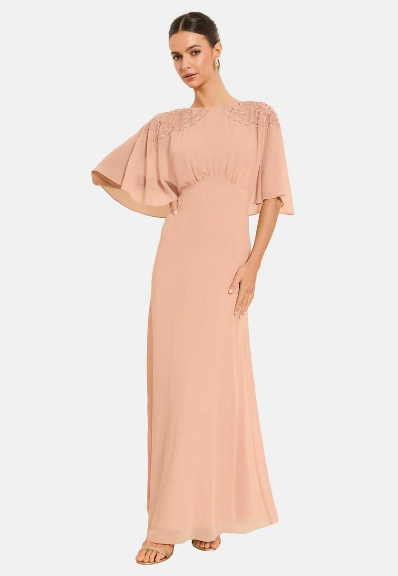 Frau trägt ein bodenlanges, roséfarbenes Kleid mit bestickten Umhangärmeln, steht mit einer Hand in der Hüfte und einem entspannten Gesichtsausdruck.