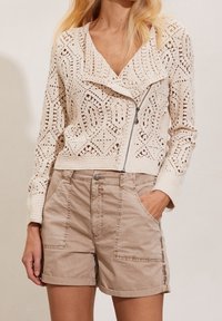 Lätt beige stickad kofta med intrikata utskurna mönster, kombinerad med bruna shorts med framfickor och uppvikta fållar.