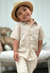 Beige linne skjort- och byxkem med kort ärm och bröstficka, tillsammans med en halmhatt. Bakgrunden innehåller en mjuk leksak och en soffa.