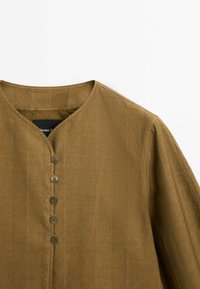 Top in tessuto leggero marrone con righe verticali; presenta un scollo a V, sette bottoni frontali e una vestibilità rilassata. Design testurizzato e informale.