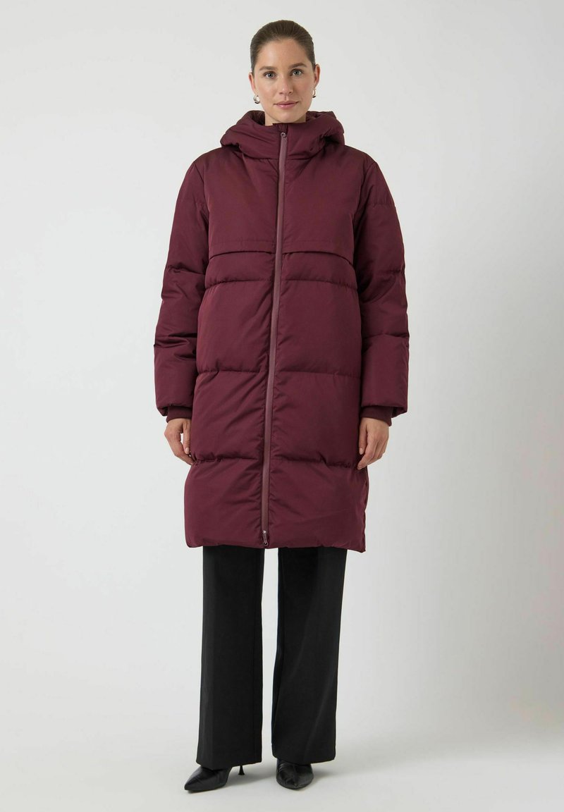 Burgunderfarget puffjakke med hette, med horisontale sømmer og hel frontglidelås. Kombinert med svarte vide bukser og høye hæler.