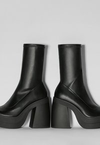 Botas negras hasta el tobillo hechas de material sintético suave, con un tacón grueso, punta redonda y un diseño sin costuras con un mínimo de costuras.