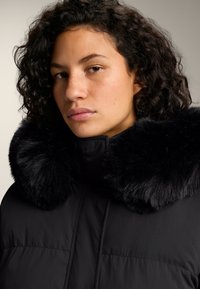 Žena s kučeravými tmavými vlasmi v čiernom zimnom kabáte s veľkým čiernym kožušinovým golierom, neutrálne vyjadrenie, svetlé pozadie.