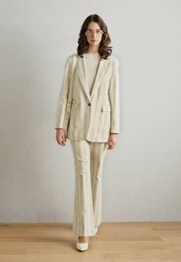 Completo a righe di colore chiaro in morbido tessuto, con giacca su misura e pantaloni svasati. Accompagnato da scarpe bianche e una borsa piccola.
