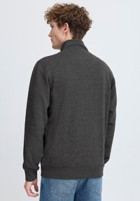 Mørk grå hettegenser med glidelås og høy krage, ribbestrikkede mansjetter og teksturert stoff. Bakfra vises et enkelt design og en avslappet passform.