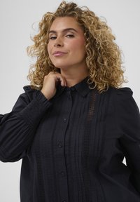 Mujer con cabello rubio rizado que lleva una blusa negra abotonada con detalles de encaje, tocándose suavemente el cuello, fondo neutro.