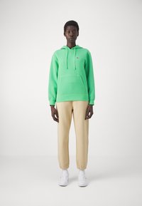 Felpa verde in cotone con tasca frontale, cappuccio con coulisse e polsini a coste, abbinata a pantaloni felpati beige e sneakers bianche.