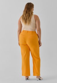 Pantaloni larghi arancioni con una texture liscia, tasche posteriori e passanti per cintura, abbinati a un top beige senza maniche e tacchi alti.