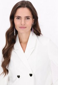 Witte double-breasted blazer met een gestructureerd ontwerp, voorzien van hartvormige zwarte knopen en een gladde stofstructuur. Lange, golvende haren en opvallende oorbellen.
