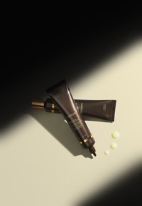 Sorte og gyldne serummoduler placeret diagonalt på en lys overflade, med tre dråber gul serum ved siden af.