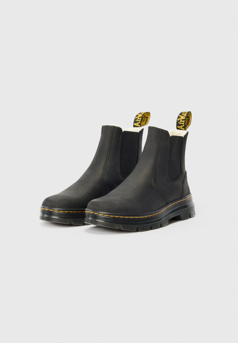 Bottes Chelsea en cuir noir avec des coutures jaunes, des panneaux élastiques sur les côtés et des tirants de marque sur un fond clair.