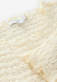 Crème-kleurige textuurstof top met gerimpelde randen en een etiket met de tekst "NEXT." Beschikt over een geweven patroon met horizontale strepen.