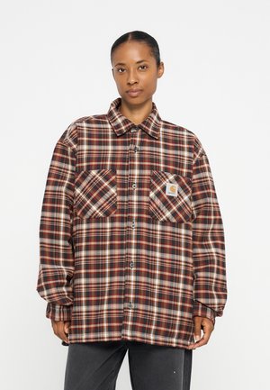 Donna che indossa una camicia a quadri marrone e rossa a maniche lunghe con due tasche sul petto e pantaloni scuri su sfondo bianco.