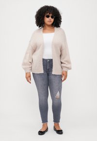 Cardigan de malha bege com frente aberta e mangas balão, usado sobre uma blusa branca e jeans cinzentos desgastados, complementado por sapatos planos pretos.