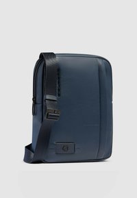 Borsa a tracolla in pelle blu navy con design piatto, tracolla regolabile e dettaglio discreto del logo in rilievo sul davanti. Texture liscia, chiusura con cerniera.