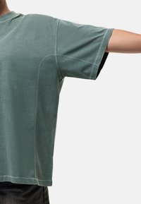 T-shirt in cotone verde chiaro con maniche corte, dal taglio ampio e dettagli di cucitura visibili. L'orlo è leggermente curvo.