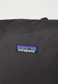Svart stoffveske med teksturert overflate og et rektangulært logetikett som viser ordet "patagonia" og fjellgrafikk i blått og rosa.