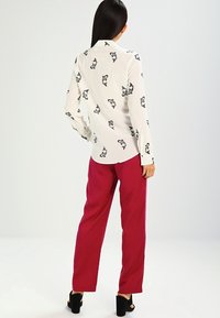 Blouse blanche à imprimé poissons noirs, manches longues et col, associée à un pantalon bordeaux taille haute et des sandales noires à talons.