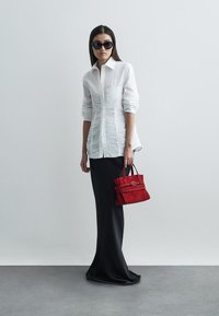 Camisa branca texturizada com botões e mangas longas, combinada com uma saia preta lisa; segurando uma pequena mala de camurça vermelha. Óculos de sol pretos.