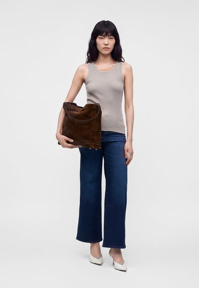 Femme portant un débardeur beige, un jean bleu à jambes larges, des talons blancs, tenant un grand sac en daim marron sur un fond clair uni.