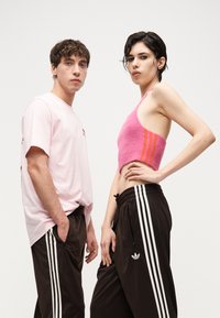 Dvaja mladí dospelí stoja vedľa seba, obaja majú čierne tepláky Adidas s bielymi pruhmi; jeden má na sebe ružové tričko, druhý ružový krátky top.