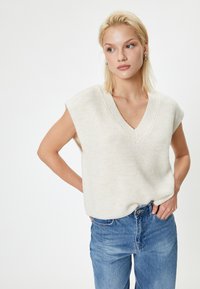 Maglione senza maniche color beige chiaro con scollatura a V, texture a costine, abbinato a jeans in denim azzurri. Design semplice e casual senza ornamenti.