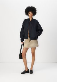 Zwarte bomberjack met een rits aan de voorkant en zijzakken, gecombineerd met een beige denim rok en zwarte instappers, terwijl ze een zwarte schoudertas vasthoudt.