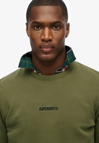Olijfgroen sweatshirt met een geribbelde kraag; eronder is een geruite shirt met een ruitpatroon in groen, rood en gele accenten. Merkl logo zichtbaar.