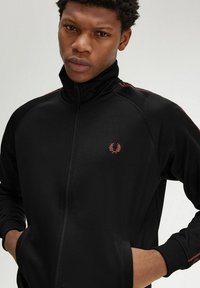 Giacca nera con zip e alto colletto, caratterizzata da strisce rosse a contrasto, polsi a coste e un piccolo logo ricamato sul lato sinistro.