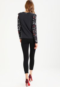 Black long-sleeve top with floral-patterned sleeves, round neckline, and small button detail at the back, paired with black leggings and red heels.