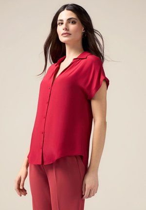 Blouse rouge à manches courtes et à boutons en tissu lisse ; présente un col et un ourlet légèrement incurvé. Associée à un pantalon rose assorti.