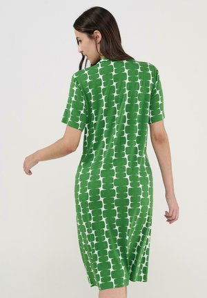 Mujer con vestido de manga corta de patrón verde y blanco hasta la rodilla, vista desde atrás contra un fondo liso.