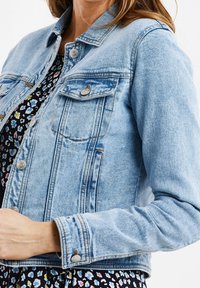 Chaqueta de mezclilla azul clara con un corte clásico, que presenta dos bolsillos en el pecho, botones plateados y detalles de costura visibles.