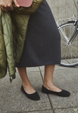 Person i sort strikket nederdel og sorte flade sko står på fortovet og holder en grøn jakke og en brun pung nær et cykelhjul.