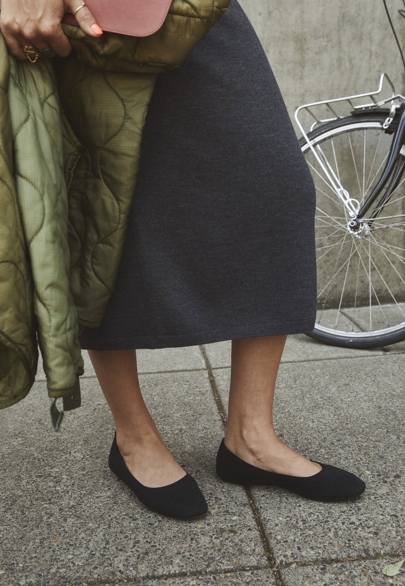Personne en jupe noire en maille et ballerines noires debout sur le trottoir tenant une veste verte et un sac marron près d'une roue de vélo.