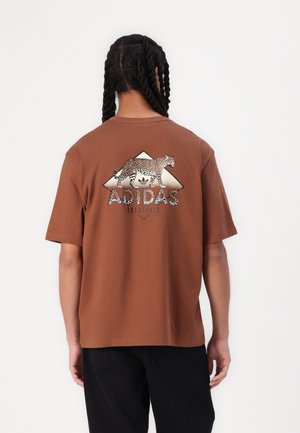 Brązowa koszulka bawełniana z grafiką lamparta i tekstem "ADIDAS ORIGINALS" w czerni, umieszczona na geometrycznym wzorze na plecach.