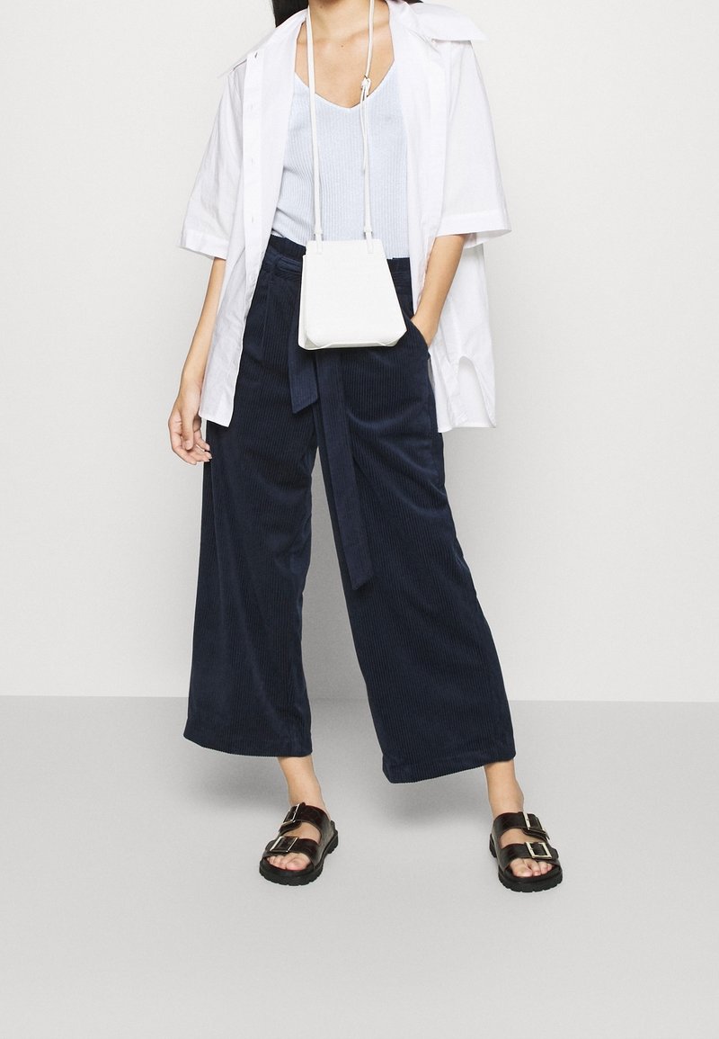 Mörkblå ribbade vida byxor med ett tygbälte, tillsammans med en ljusblå linne, en vit oversized skjorta och svarta sandaler med spännen.