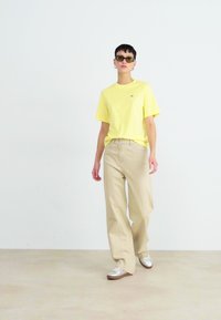 Camiseta de algodón amarilla combinada con pantalones de pierna ancha beige y zapatillas plateadas. La camiseta presenta un pequeño logotipo cerca del escote.