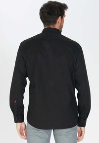 Chemise noire à manches longues avec des poignets boutonnés et un détail logo rouge discret sur la manche gauche. Elle présente un col et un design structuré.