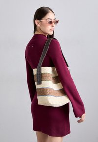 Burgundi színű ruhát viselő nő, rózsaszín napszemüveget hord, vállán egy sötétbarna fogantyúkkal rendelkező, szőtt csíkos táskával.