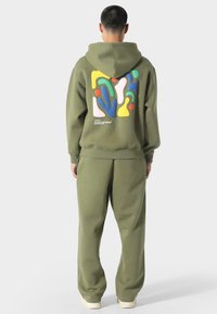 Oliven grøn hoodie og sweatpants sæt, der har et farverigt abstrakt tryk på ryggen. Fremstillet af blødt fleece-materiale.