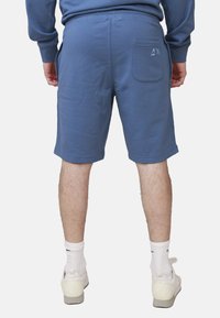 Pantaloni corti blu in cotone con vita elasticizzata, tasche laterali e una tasca posteriore con un piccolo logo. Indossati con sneakers bianche e calze.