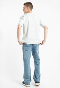 Młody mężczyzna w białej koszulce, niebieskich dżinsach i czarnych sneakersach stoi, odwrócony plecami, trzymając obiema rękami tył swojej koszulki.