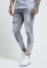 Grå denim slim jeans med slitna detaljer och revor, kombinerat med vita träningsskor. Slät textur med en åtsittande silhuett.